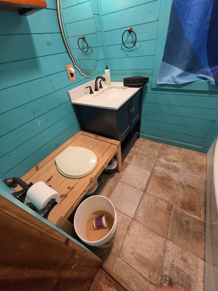 A composting toilet: a wooden bench with a seat, beside a bucket of sawdust, in a teal-walled bathroom.