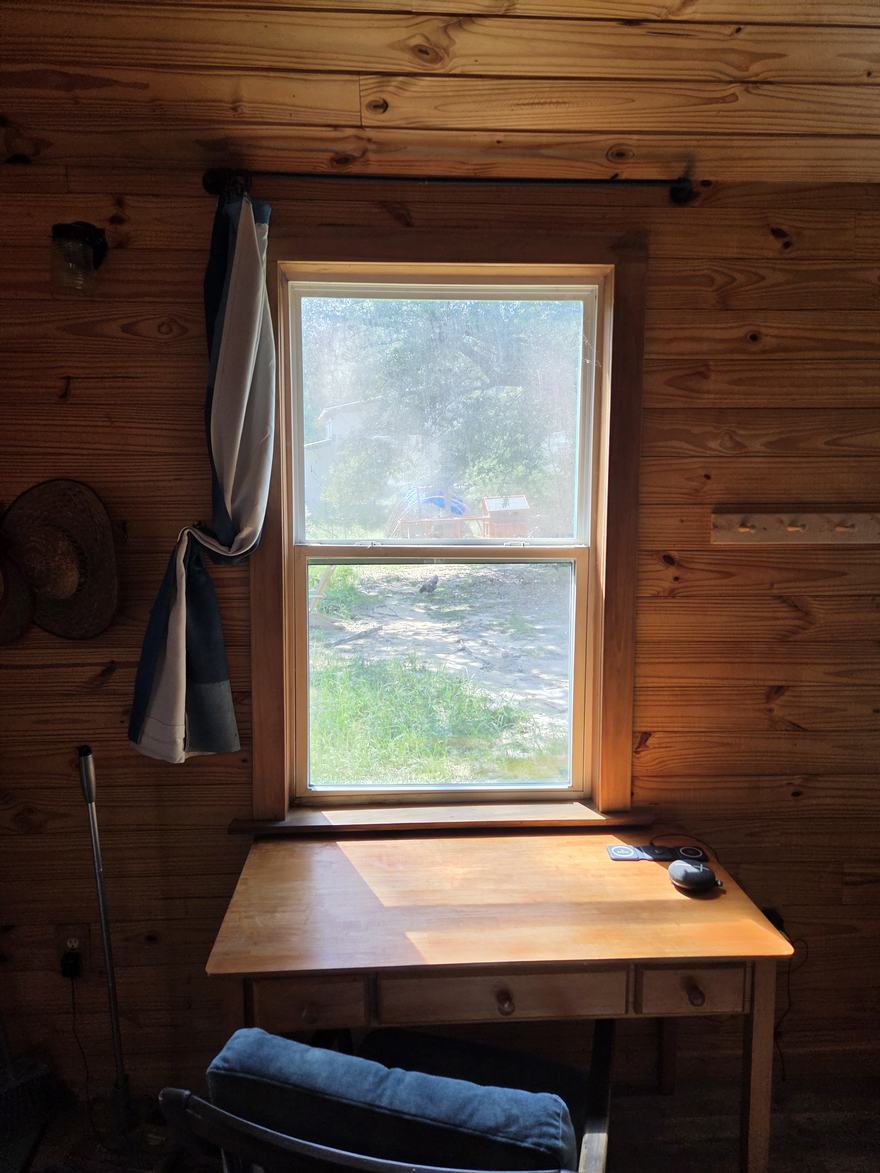 A small wooden desk beneath a cabin window, afternoon light falling across it, a straw hat on the wall nearby.