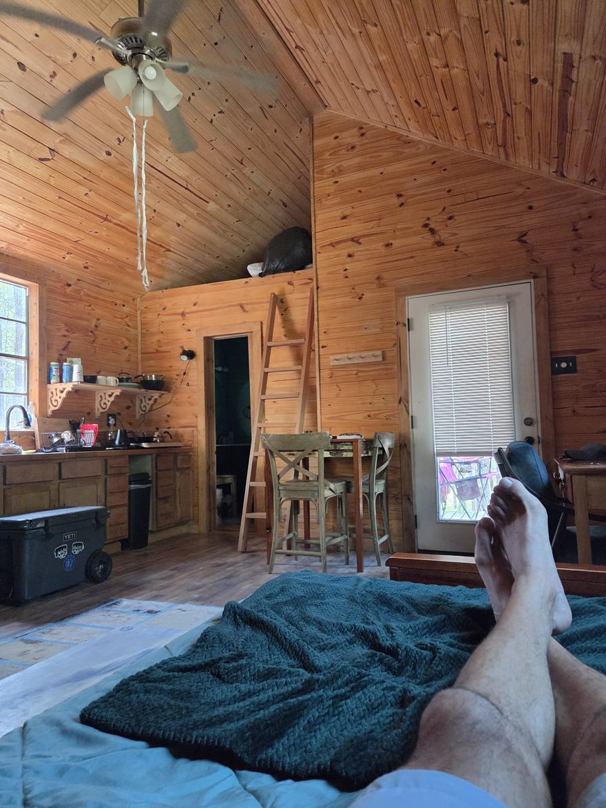 View from the bed across the cabin: feet up on the blanket, ceiling fan turning, ladder to the loft, front door beyond.