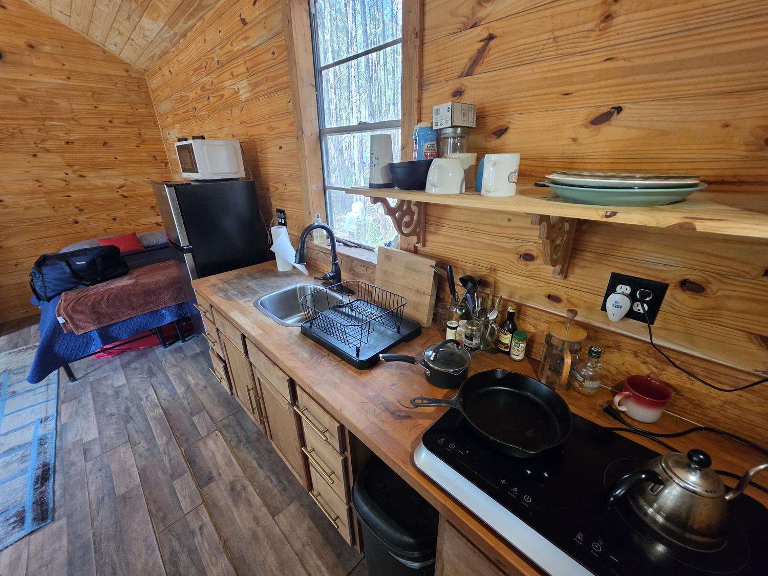 The cabin kitchen counter: butcher block, a cast iron skillet on a portable induction burner, kettle, dish rack, and a shelf of mismatched mugs and plates.